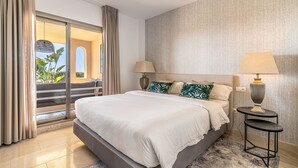 A modern bedroom with a large bed, two bedside tables with lamps, a view through a sliding door to an outdoor area, and a textured wall. - Premium 2BR with Beautiful Terrace & Sea Views (Benahavís)