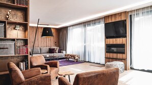 A modern living room with a sofa, armchairs, a coffee table, a bookshelf, a TV, and a large window with curtains. - Chalet Amadeus - Designer Luxury Chalet for 14 (Piesendorf)