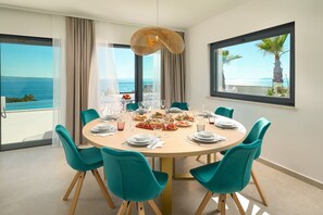Ein Esszimmer mit einem runden Tisch, gedeckt für sechs Personen, großen Fenstern mit Meerblick, blaugrünen Stühlen und einer hängenden Leuchte.