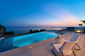 Una piscina infinita moderna con vista despejada al océano, un área de descanso con una silla y una pequeña mesa con una botella y una copa.