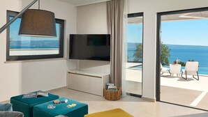 A modern living room with a flat-screen TV, teal and mustard ottomans, a grey sofa, and a view of the sea through large glass doors. - Villa Sunset - Luxury Adriatic Sea View Estate (Podstrana)