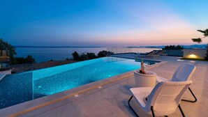 Una piscina infinita moderna con vista despejada al océano, un área de descanso con una silla y una pequeña mesa con una botella y una copa.