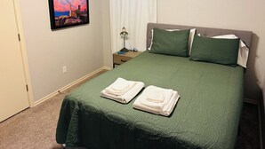Un lit bien fait, avec une literie verte et deux serviettes blanches pliées, une lampe de chevet et, au mur, un tableau représentant un paysage urbain encadré.
