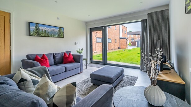 A modern living room with a grey sofa, red cushions, a grey ottoman, and a large window with a view of a brick building and a green lawn. - Garden View - sleeps 6 guests  in 2 bedrooms (York)