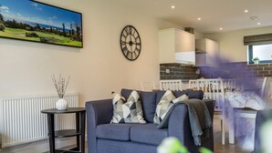 A living room with a grey sofa, a black clock, a white radiator, and a dining area with a white table and chairs. - Garden View - sleeps 6 guests  in 2 bedrooms (York)