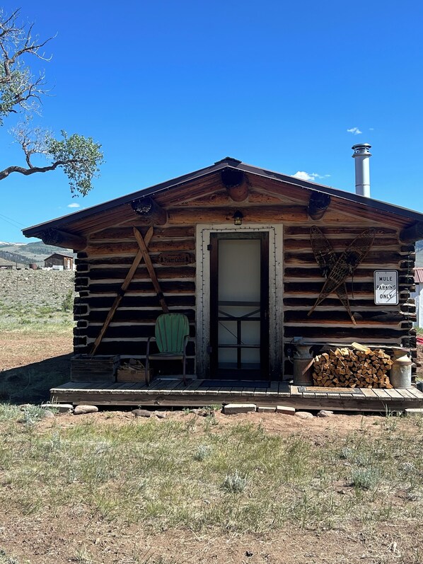 Un chalet rustique en rondins de bois, avec une porte en bois, une chaise verte à l’extérieur et un panneau indiquant : « Stationnement réservé aux mulets ».