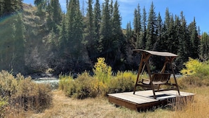 A wooden swing chair on a wooden platform in a forested area with a river in the background. - Rel Rio Rentals (Creede)