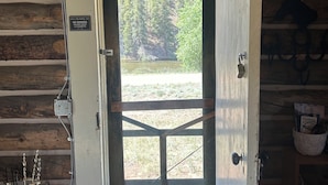 A wooden cabin with a door leading to a view of a forest and a body of water. - Rel Rio Rentals (Creede)