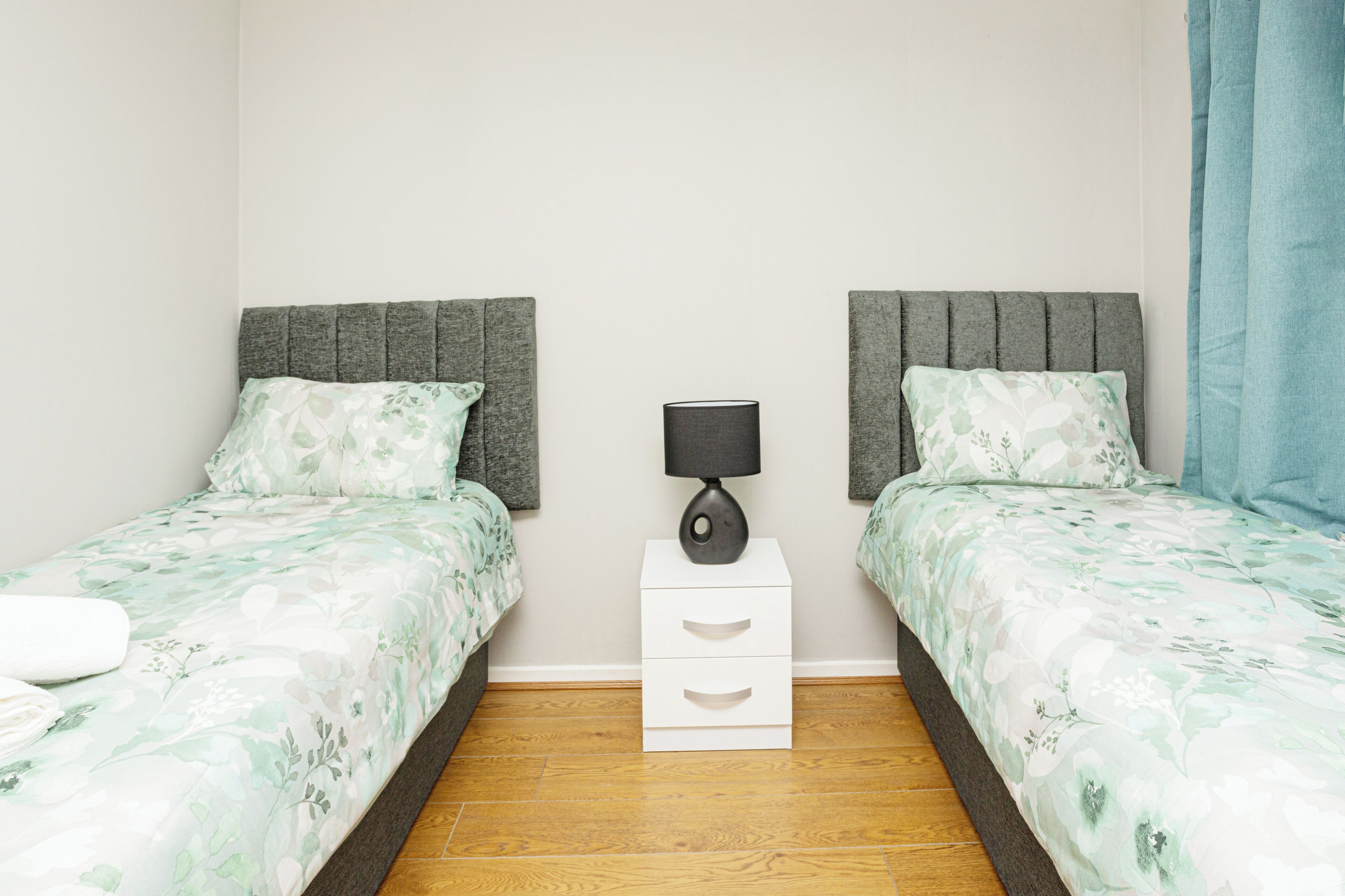 Une chambre avec deux lits, chacun doté d’une tête de lit grise et d’une literie à motifs floraux. Une table de chevet blanche avec une lampe se trouve entre les deux lits.