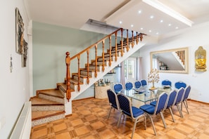 A dining area with a glass table and blue chairs, a staircase with wooden balusters, and a Buddha statue on the wall.