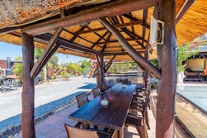 An outdoor wooden pavilion with a thatched roof, surrounded by a gravel area with a pool and lounge chairs.