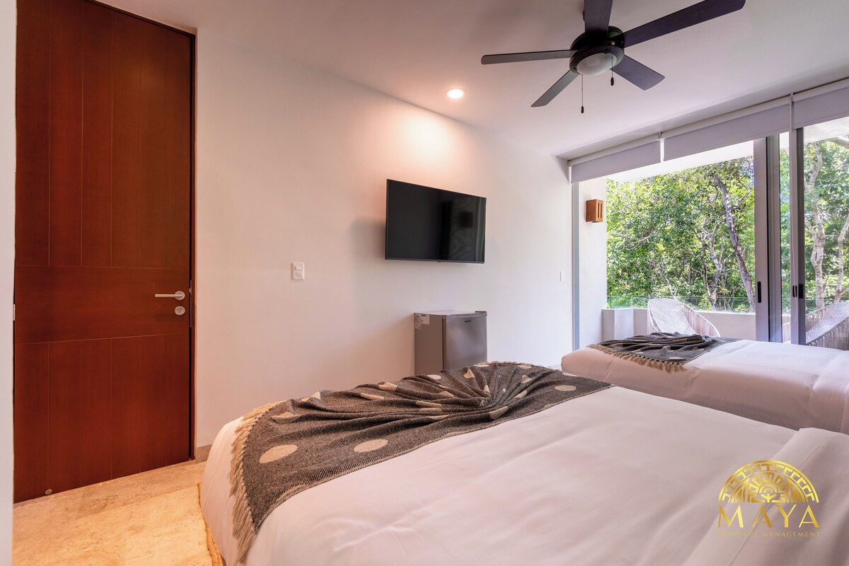 A hotel room with two beds, a ceiling fan, a TV mounted on the wall, and a large window offering a view of trees.