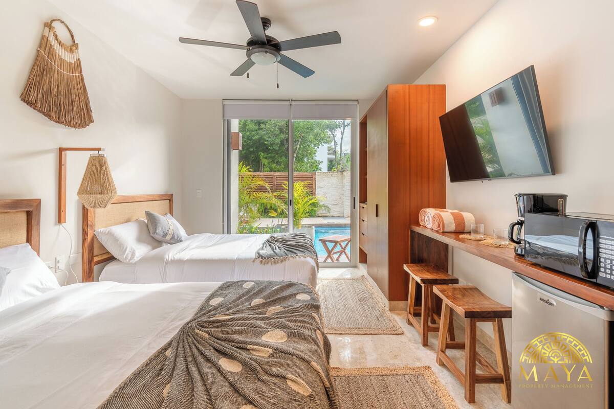Quarto de hotel com duas camas, ventoinha no teto, televisão, micro-ondas e vista para uma piscina e vegetação.