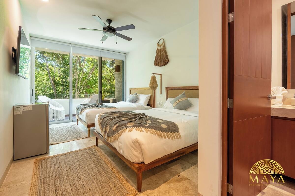 Quarto com duas camas, ventilador de teto, geladeira e porta de vidro corrediça para uma varanda.