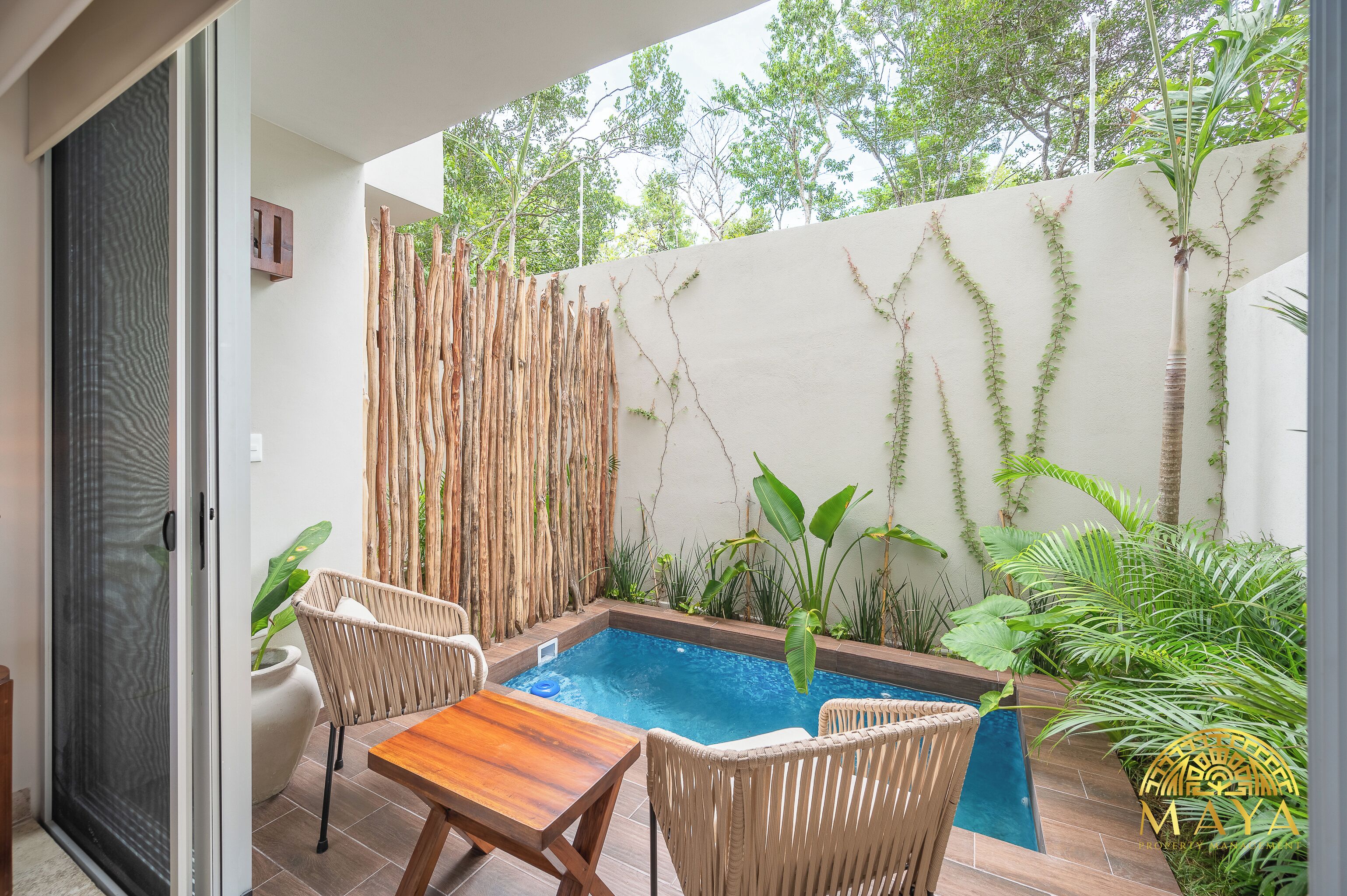 A small outdoor pool area with a wooden table and wicker chairs, surrounded by lush greenery and a bamboo fence.