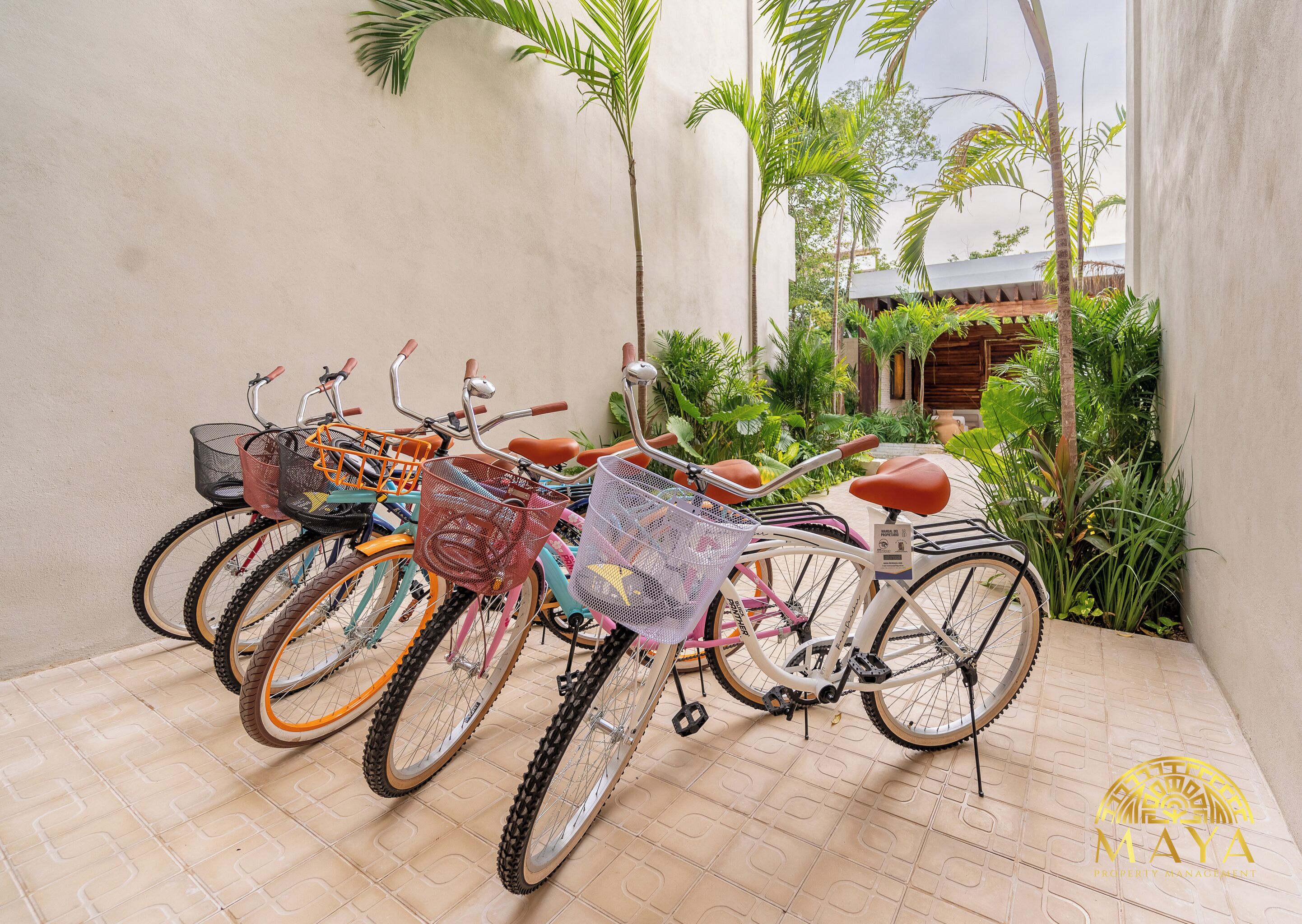 Bicicletas estacionadas em área externa com piso de azulejos, cercadas por vegetação e um edifício ao fundo.