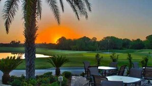 Un espace extérieur aménagé avec des tables et des chaises, offrant une vue sur un terrain de golf et un plan d’eau au coucher du soleil.