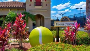 Un court de tennis orné d’une grande sculpture représentant une balle de tennis, entouré de plantes tropicales et bordé d’un bâtiment sur lequel est apposée une enseigne indiquant la présence d’un court de tennis.