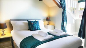 Una cama bien hecha con sábanas blancas y un plaid color turquesa, una lámpara de noche y una mesita con una lámpara.