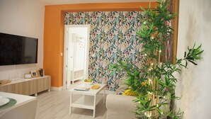A living room with a flat-screen TV, a white coffee table with a bowl of fruit, a sofa, and a large window with a view of the outdoors. - Apartment in the center of Sanlúcar (Sanlúcar de Barrameda)