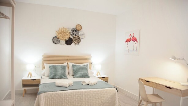 A modern bedroom with a large bed, two bedside tables with lamps, a desk with a chair, and a wall art piece of flamingos. - Apartment in the center of Sanlúcar (Sanlúcar de Barrameda)