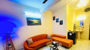 A modern living room with a curved orange sofa, a glass coffee table, and a small black cabinet. There is a painting on the wall and a chandelier above the sink. - Cafe Green Valley (Rangpur)