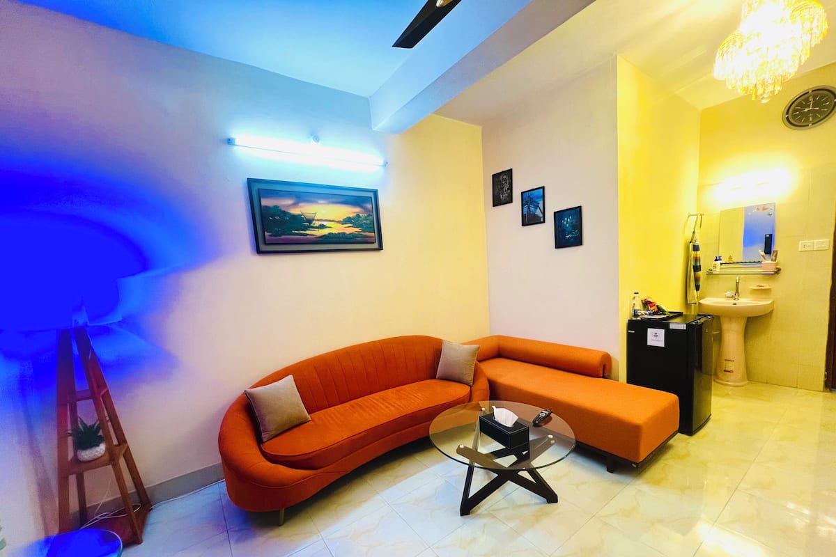A modern living room with a curved orange sofa, a glass coffee table, and a small black cabinet. There is a painting on the wall and a chandelier above the sink.