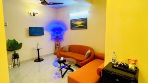 A living room with a sofa, a glass coffee table, a small black table, a ceiling fan, a wall-mounted TV, and a framed picture. - Cafe Green Valley (Rangpur)