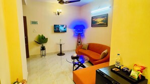 A living room with a sofa, a coffee table, a TV, a ceiling fan, and a refrigerator. - Cafe Green Valley (Rangpur)