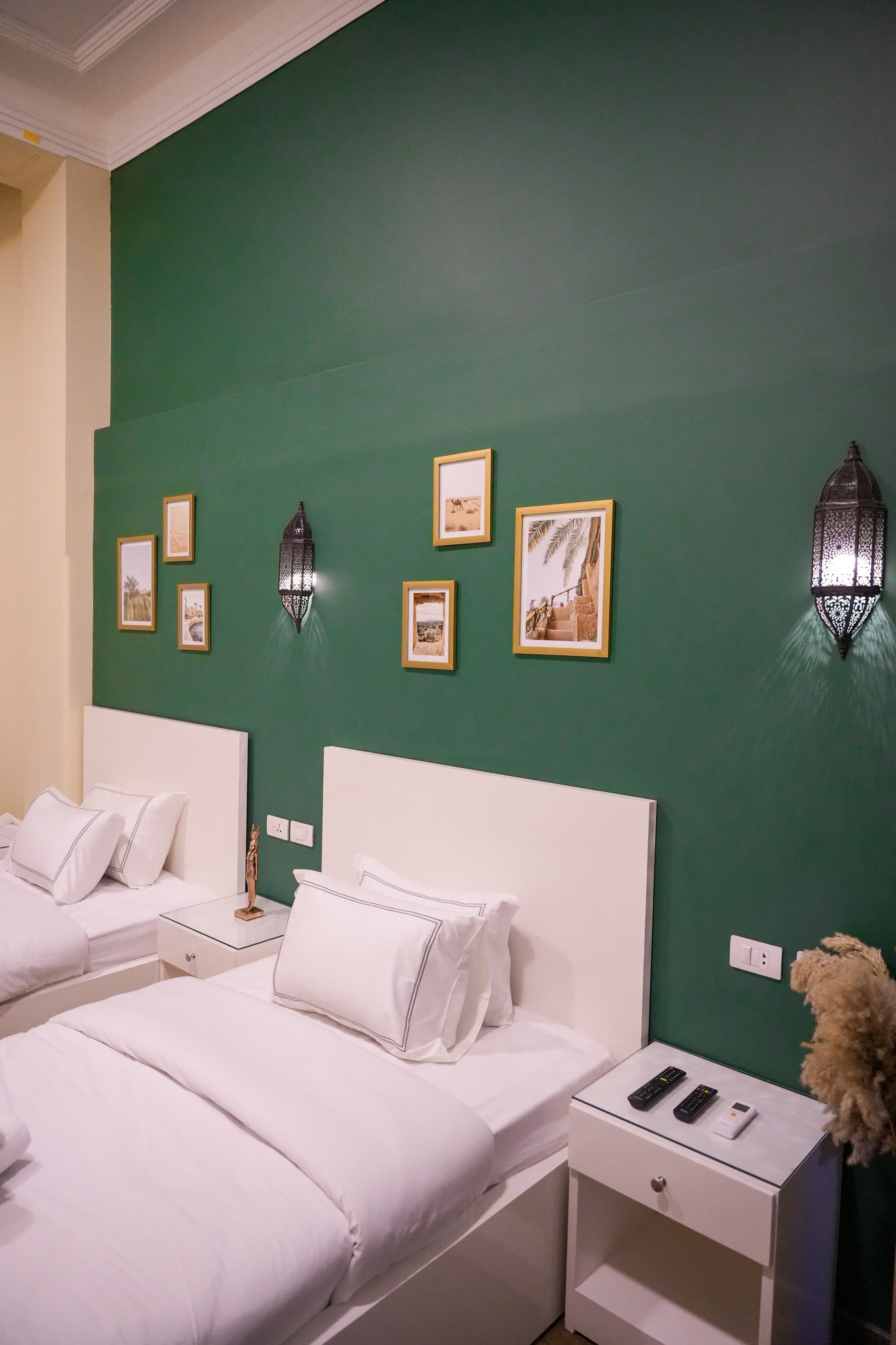 A hotel room with two beds, a green accent wall, framed pictures, a decorative lantern, and a nightstand with a remote control.