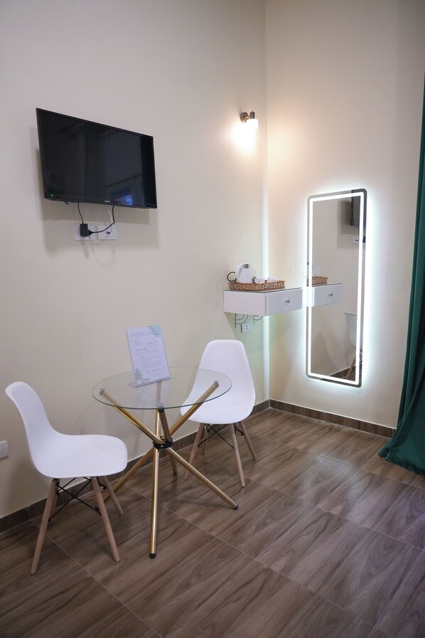 Una habitación pequeña con una mesa redonda de vidrio, dos sillas blancas, un televisor de pantalla plana, un espejo con luces y un estante con artículos de aseo.