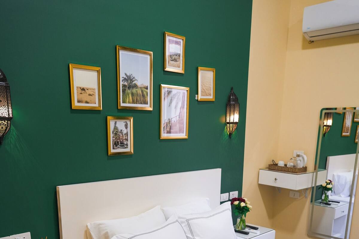 Una cama bien hecha con sábanas blancas, un plaid verde y una bata sobre ella. Hay cuadros en la pared, una mesita de noche con una lámpara y una pequeña estatua.