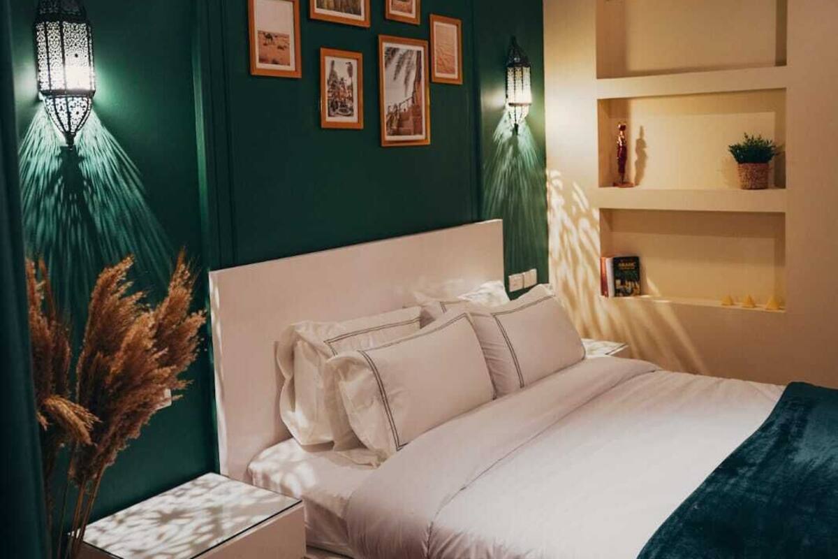 Un dormitorio con cama blanca, dos mesitas de noche, una mesita auxiliar, una estantería de pared y dos lámparas de pared.