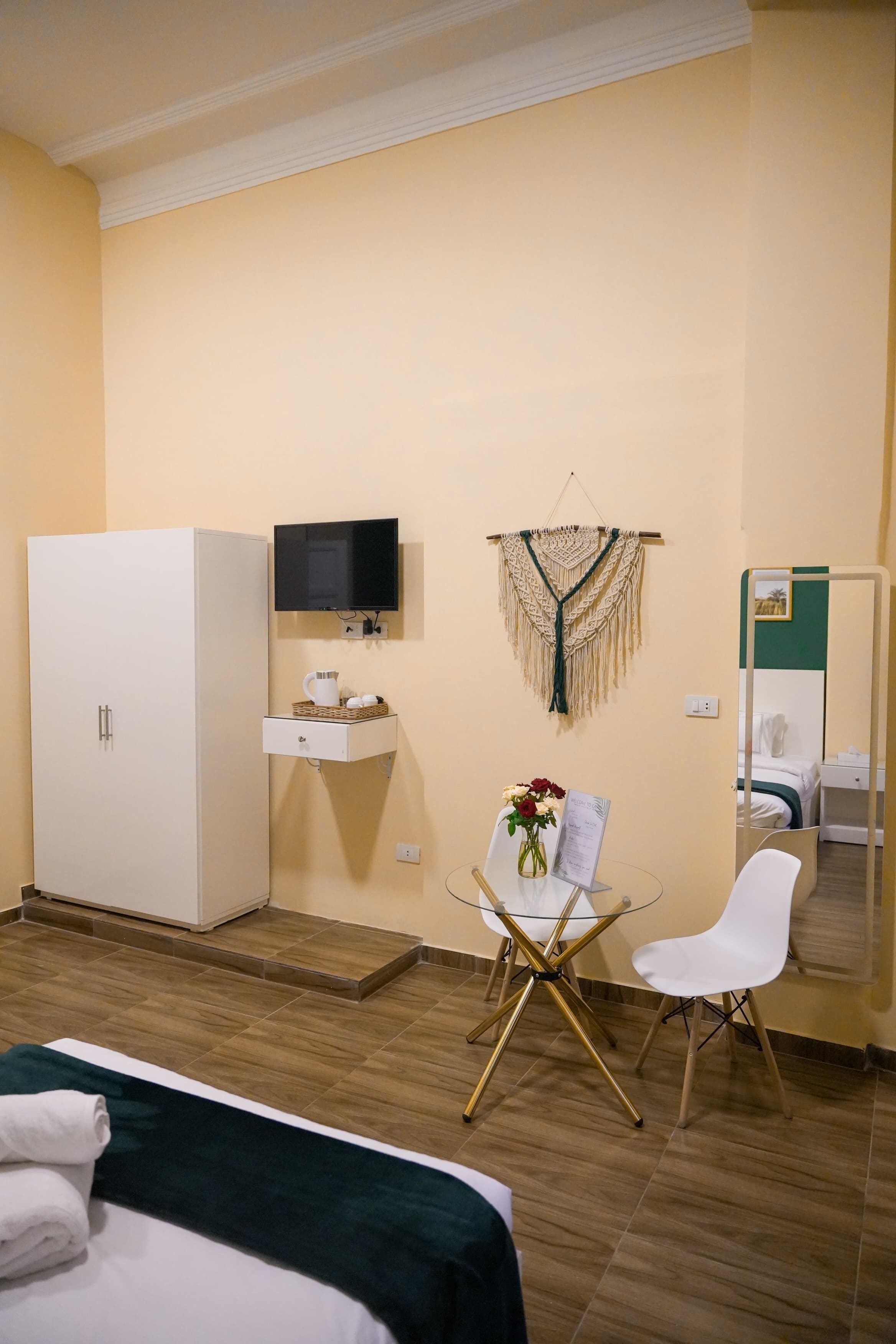 Ein Hotelzimmer mit Bett, einem weißen Schrank, einem kleinen Tisch mit Blumenvase, einem Sessel, einem an der Wand montierten Fernseher und einem Spiegel.