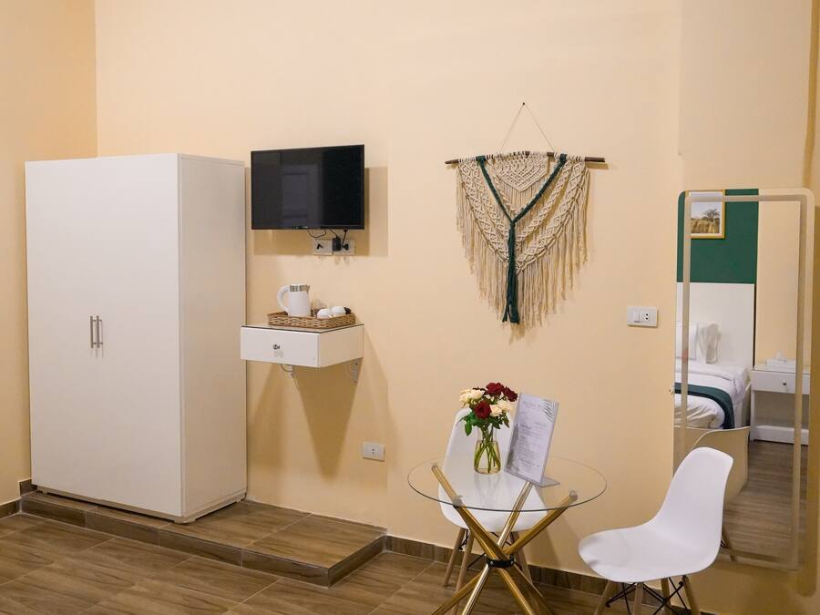 Ein Hotelzimmer mit Bett, einem weißen Schrank, einem kleinen Tisch mit Blumenvase, einem Sessel, einem an der Wand montierten Fernseher und einem Spiegel.