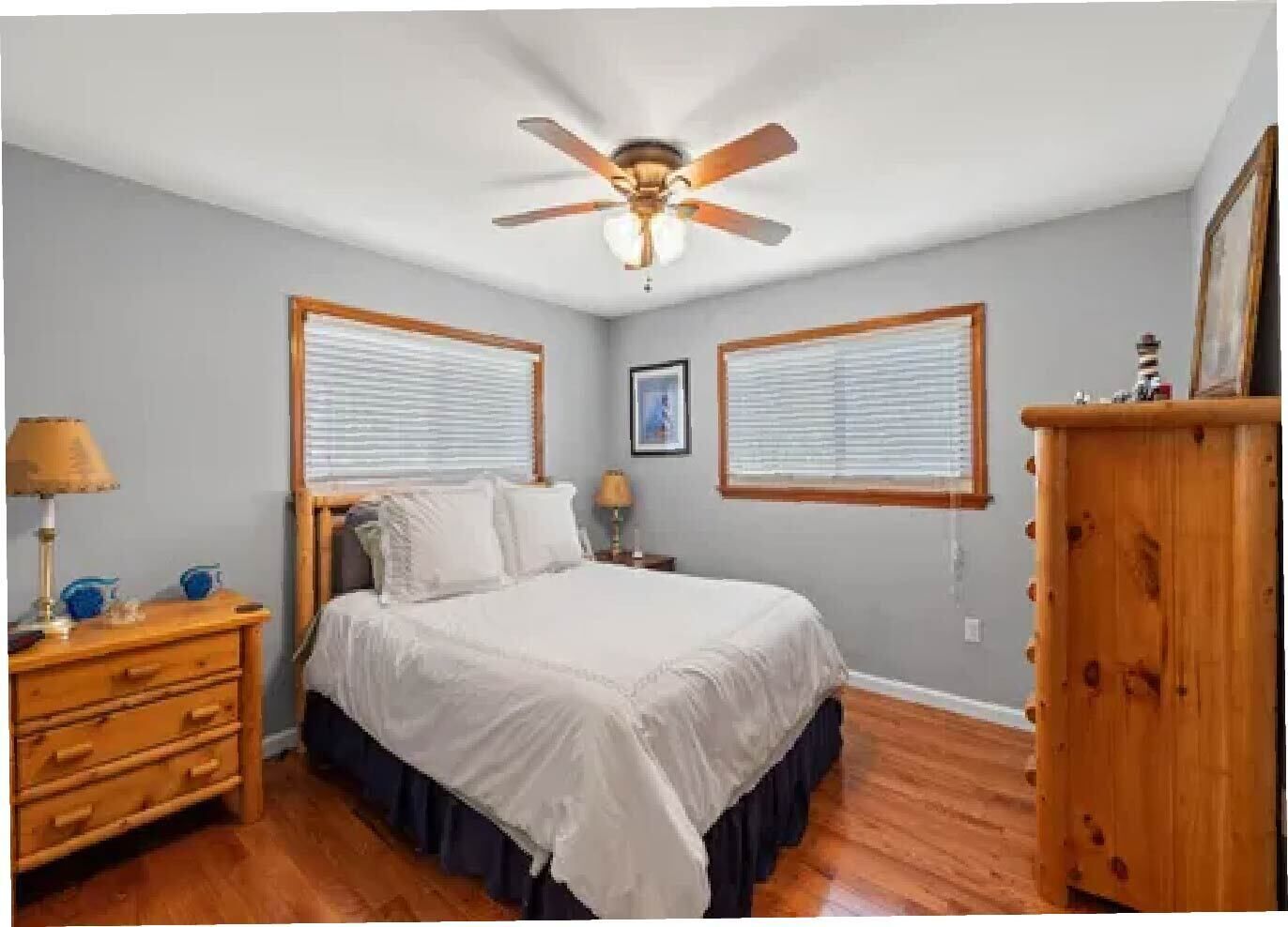 A bedroom with a bed, two bedside tables, a dresser, a ceiling fan, and two windows with blinds.