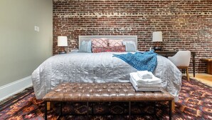 Een slaapkamer met een bakstenen muur, een bed met beddengoed met een patroon, een bankje en een tapijt met dessin.
