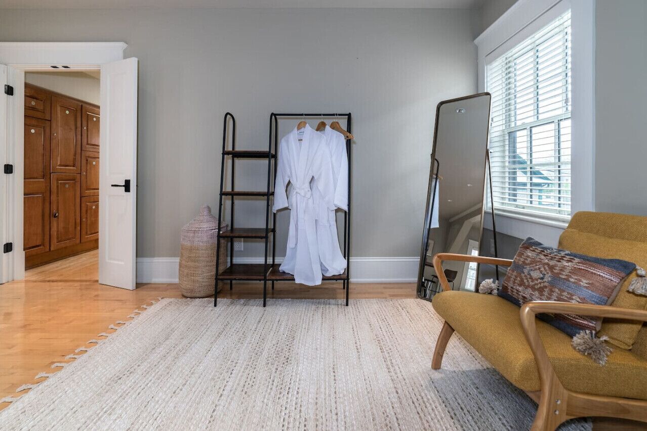 Una habitación con un armario de madera, una bata blanca sobre un perchero, un sillón amarillo y un ventanal con persianas.