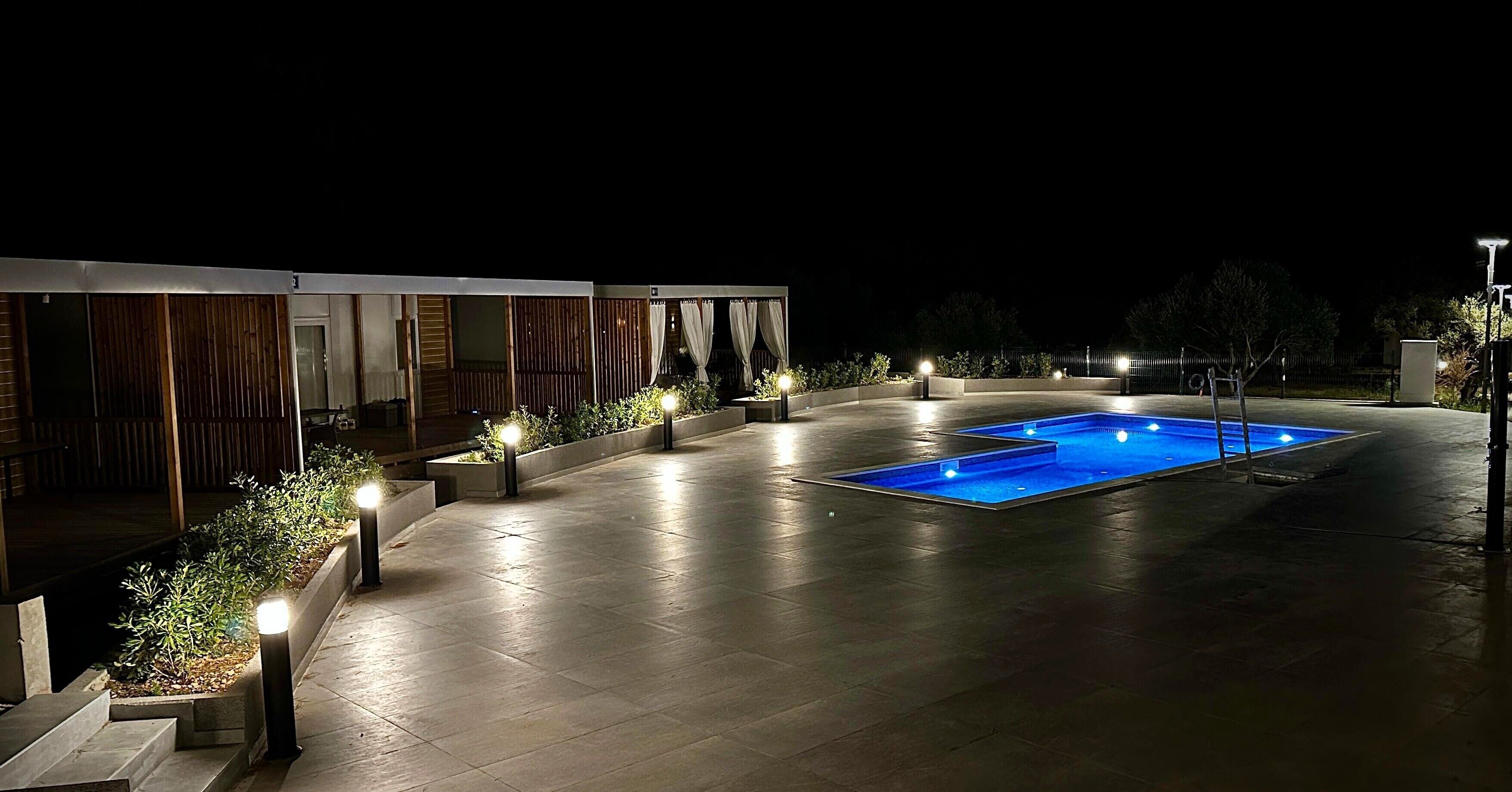 Un espace piscine extérieur moderne, doté d’une piscine rectangulaire, entourée d’une terrasse dallée et éclairée par des luminaires encastrés dans le sol.