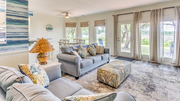 A spacious living room with a grey sofa set, a patterned ottoman, and a lamp. Large windows with curtains offer views of the outdoors. - Walk to Marina: Pet-Friendly Little River Home! (Little River)