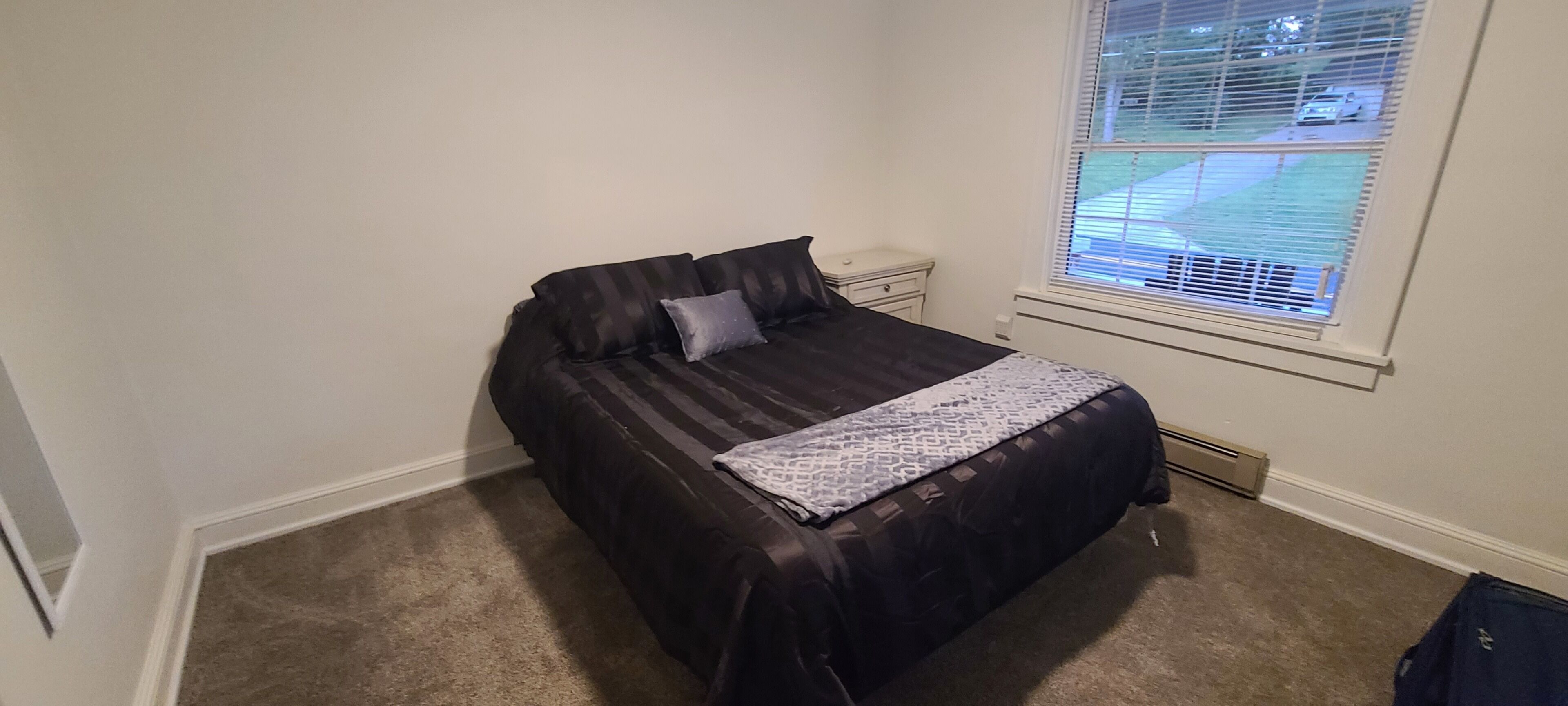 A bedroom with a bed, a nightstand, a window with blinds, and a carpeted floor.