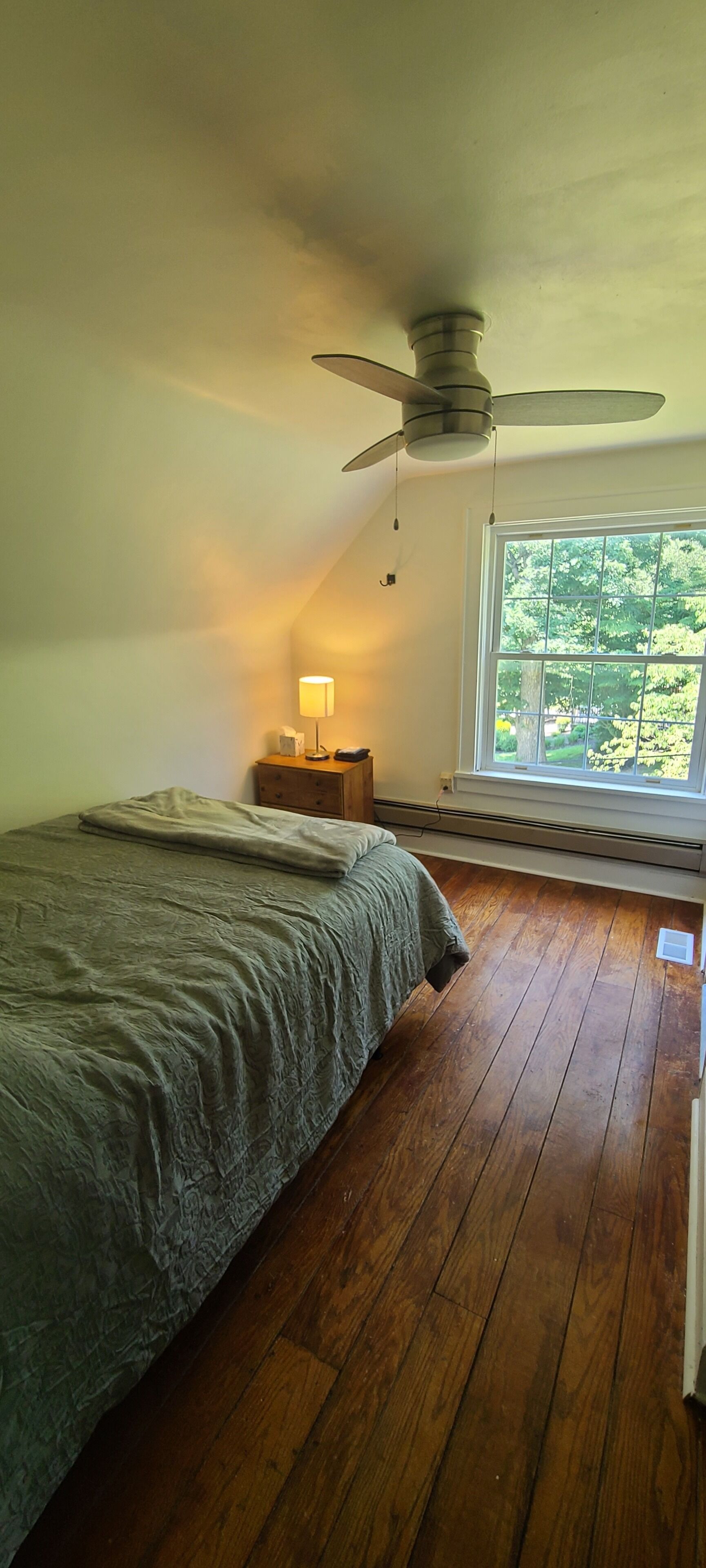A bedroom with a bed, a nightstand, a lamp, a ceiling fan, and a window with a view of greenery.