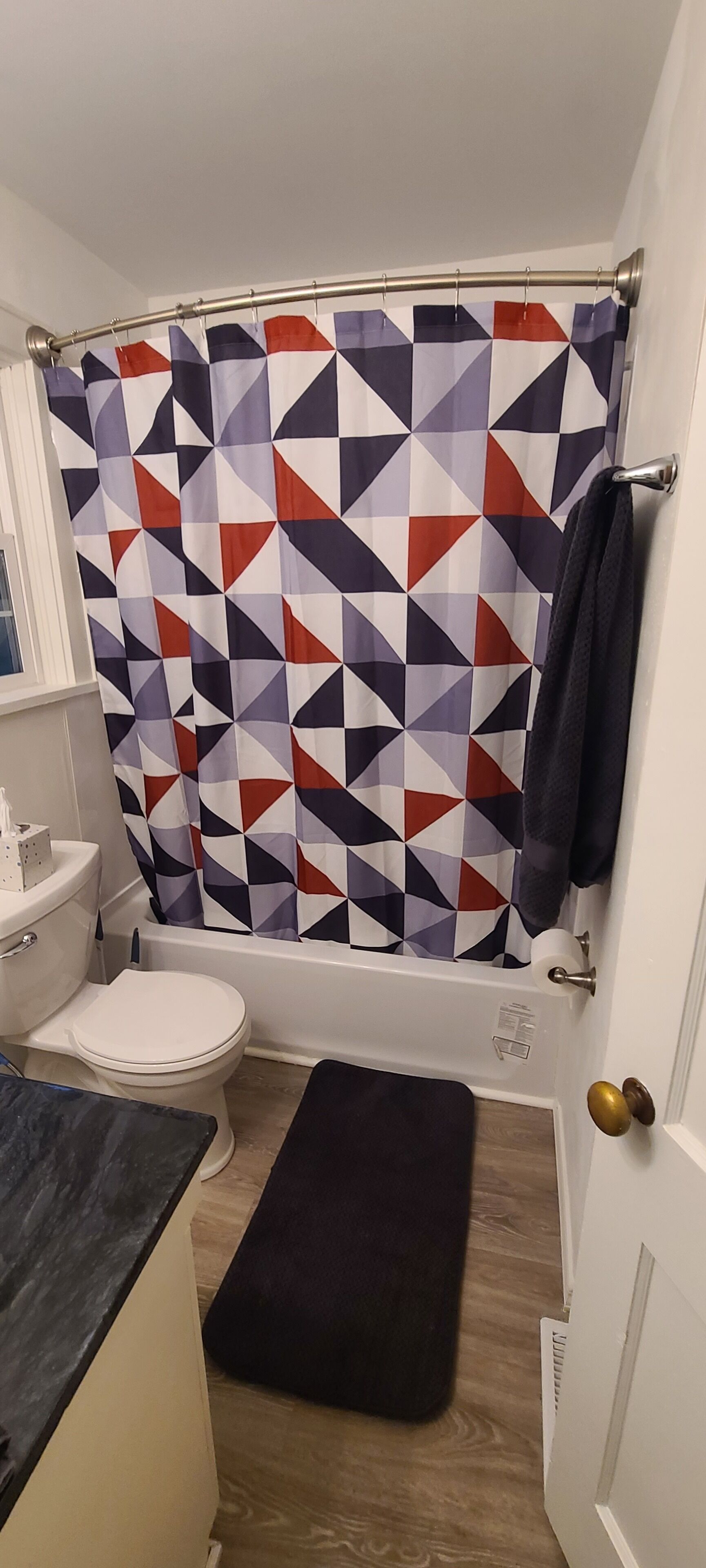 Un bagno con tenda da doccia a motivi geometrici, un water bianco, un tappetino da bagno nero e un asciugamano appeso alla porta.