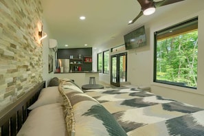 Een slaapkamer met een groot bed, een stenen muur, een televisie, een kitchenette en een raam met uitzicht op bomen.