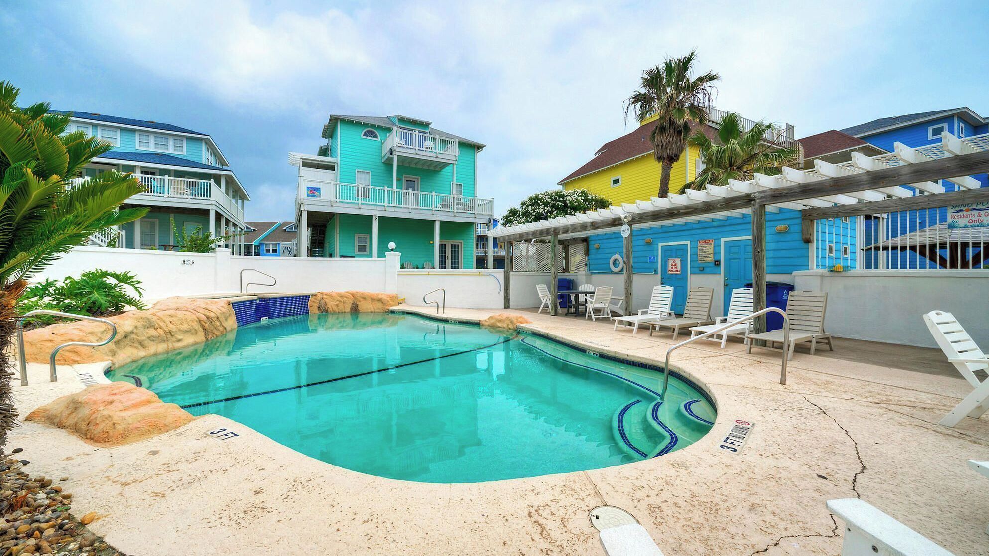 A swimming pool with a sandy edge, lounge chairs, and a building with a blue facade.