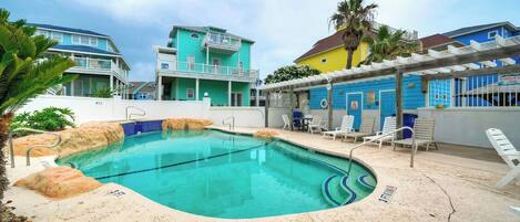 A swimming pool with a sandy edge, lounge chairs, and a building with a blue facade.