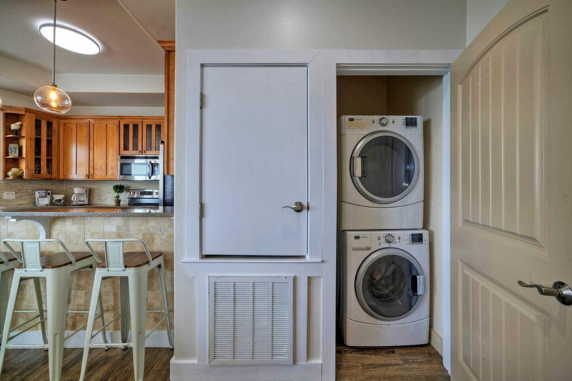 Un cuarto de lavandería con lavadoras y secadoras apiladas, una puerta, y una kitchenette con gabinetes de madera y una lámpara colgante.