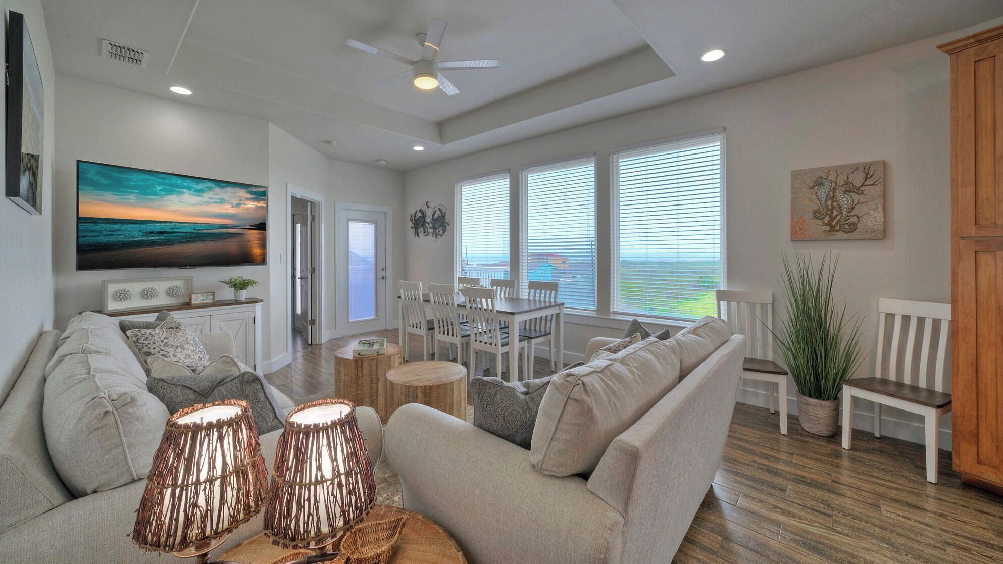 A spacious living room with a large flat-screen TV, a dining area with white furniture, and a view of the ocean through large windows.