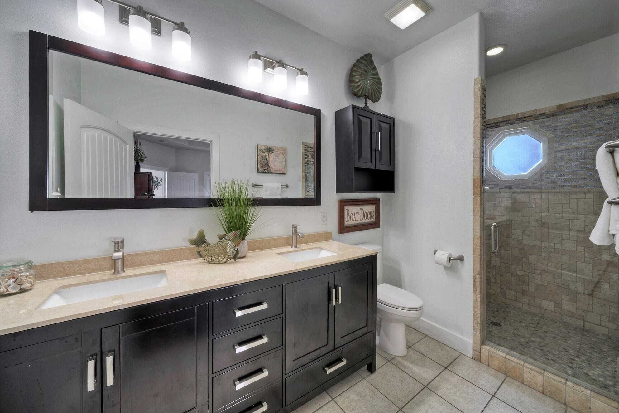 A bathroom with a large mirror, two sinks, dark cabinets, a toilet, and a glass shower enclosure.