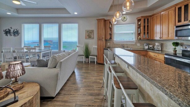 A spacious living area with a sofa, dining table, and chairs, a kitchen with wooden cabinets and a large island, and a view through the windows.